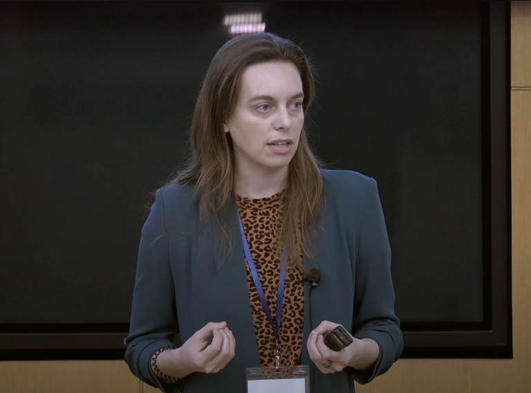 A woman wearing business attire and holding a presentation clicker speaks.